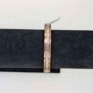 May include: A copper cuff bracelet with a silver geometric pattern. The bracelet is displayed on a black velvet surface against a white background. The cuff is approximately 0.75 inches wide.
