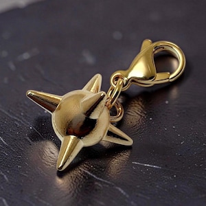 May include: A gold-toned metal charm featuring a spherical center with four protruding spikes, attached to a lobster claw clasp. The charm has a polished, reflective surface and is designed to be clipped onto jewelry or other accessories.