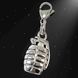 May include: A silver-toned metal charm shaped like a hand grenade, featuring a textured, grid-like surface pattern and a top lever mechanism. The charm is attached to a small, functional silver-colored lobster claw clasp by a circular jump ring.