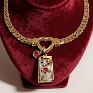 May include: A gold-toned chain necklace featuring a heart-shaped clasp and a rectangular pendant depicting a tarot card design labeled THE LOVERS. The pendant includes a red heart, a crescent moon, and small clear stones, accompanied by a small red charm.