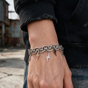 Könnte beinhalten: Ein silberfarbenes Gliederkettenarmband mit mehreren hängenden Blitz-Anhängern. Das Armband besteht aus ineinandergreifenden kreisförmigen Metallgliedern, an denen in Abständen mehrere kleine, flache Blitz-Anhänger befestigt sind.