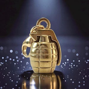 May include: A gold-toned metal charm shaped like a hand grenade, featuring a textured grid pattern on the body, a lever mechanism on the side, and a circular jump ring at the top for attachment to a chain or bracelet, with a polished finish.