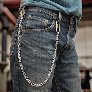 Puede incluir: Una cadena de metal plateado para cartera con un complejo patrón de eslabones entrelazados. La cadena está sujeta a unos vaqueros de mezclilla azul con un mosquetón plateado y un gancho de metal, creando un efecto drapeado en la pierna.