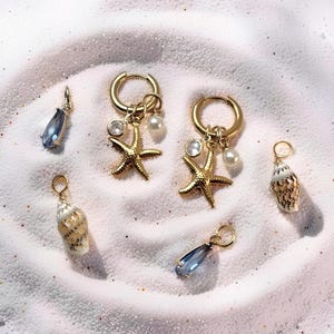 May include: Gold-toned hoop earrings with starfish, pearl, and crystal charms. Additional charms include teardrop-shaped blue gems and small seashells. The jewelry is displayed on a white, sandy surface.