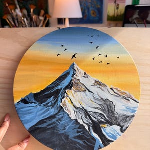 May include: A circular canvas painting depicting a snow-capped mountain peak against a vibrant sunset sky. The scene features deep blue shadows on the mountain slopes, bright white highlights on the summit, and a flock of black birds flying above.
