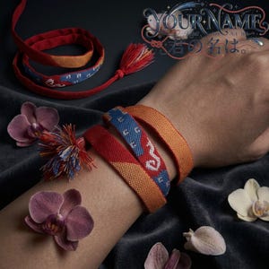 Puede incluir: Una pulsera de tela tejida con un patrón multicolor de naranja, rojo y azul con detalles geométricos blancos. La pulsera incluye una borla decorativa y está envuelta alrededor de una muñeca. El texto "YOUR NAME" es visible en la esquina.