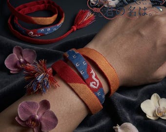 May include: A woven fabric bracelet featuring a multi-colored pattern of orange, red, and blue with white geometric accents. The bracelet includes a decorative tassel and is wrapped around a wrist. The text "YOUR NAME" is visible in the upper corner.