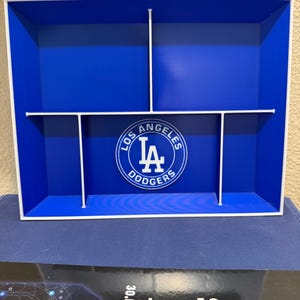 Könnte beinhalten: Ein rechteckiges blaues Aufbewahrungstablett mit weißen Trennwänden, die fünf separate Fächer bilden. In der Mitte des Tabletts befindet sich ein kreisförmiges, weiß-blaues Logo der Los Angeles Dodgers. Das Tablett hat einen weißen Rahmen.