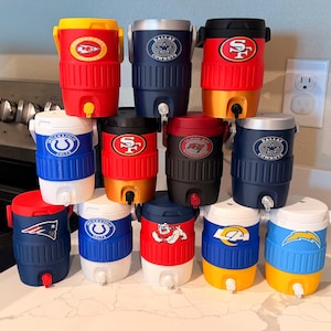 May include: A collection of twelve plastic beverage coolers arranged in a pyramid, each featuring a unique NFL team logo. The coolers come in various colors including red, blue, black, white, and yellow, and each includes a white or black spigot.