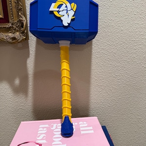 Puede incluir: Un martillo azul con mango amarillo y el logotipo blanco y amarillo de los Los Angeles Rams en la cabeza. El martillo descansa sobre un libro rosa con el texto all good taste y kate spade new york impreso en la portada en letras blancas.