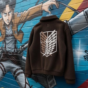 May include: A brown fleece jacket with a white and brown emblem, hanging on a hanger in front of a colorful mural. The mural features an anime character and the words "SCOUT REGIMENT".