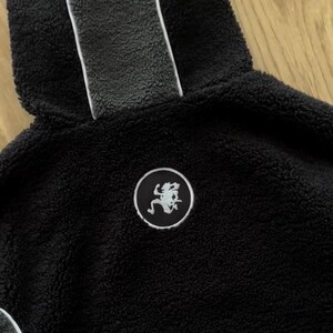 May include: A close-up of a black hooded sweatshirt made from a plush, textured material. The hood has a grey stripe down the middle with a white border. A circular patch with a white graphic is on the chest. A warm outerwear garment.