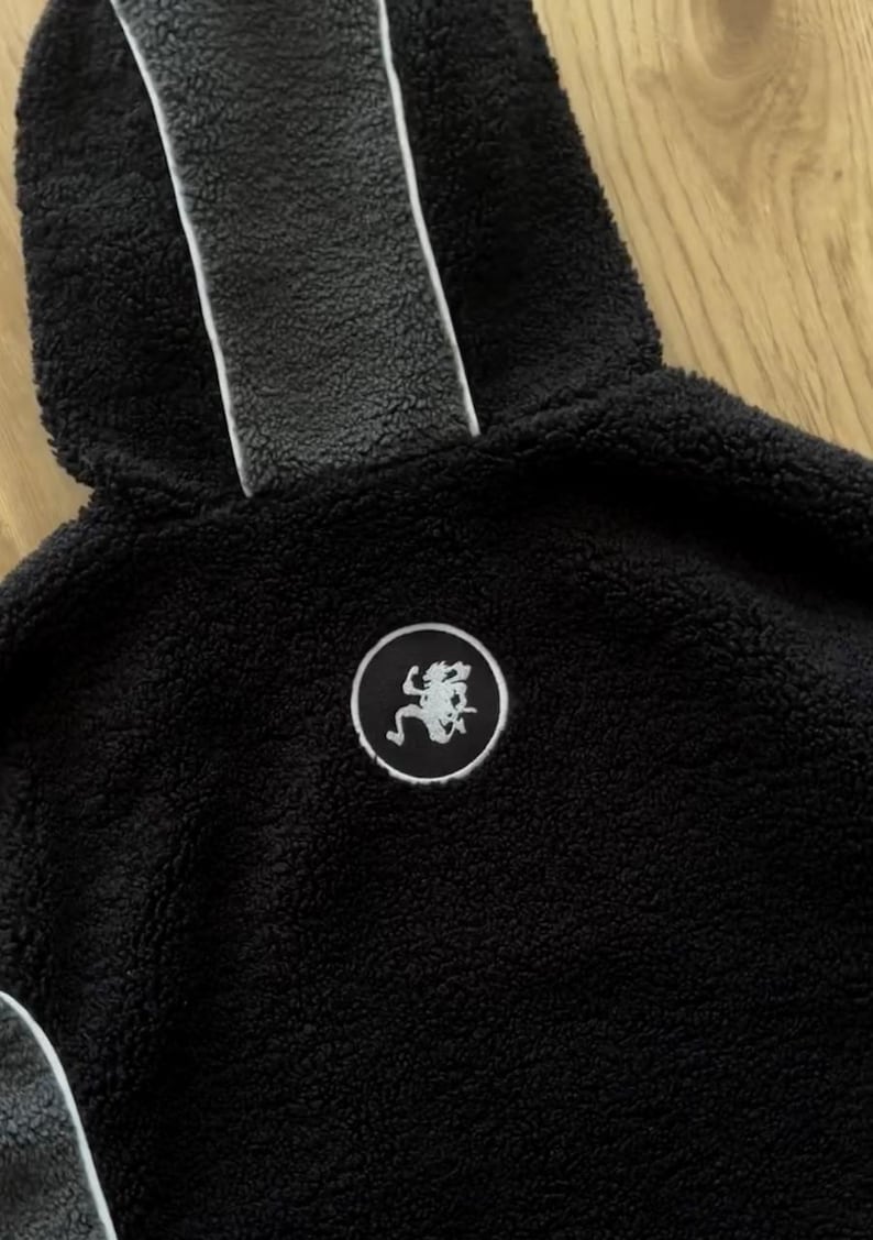 May include: A close-up of a black hooded sweatshirt made from a plush, textured material. The hood has a grey stripe down the middle with a white border. A circular patch with a white graphic is on the chest. A warm outerwear garment.