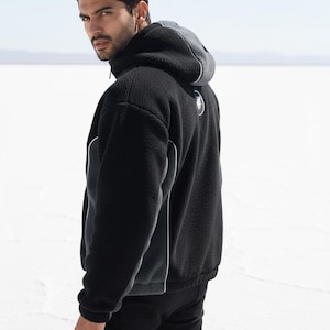May include: Black fleece hooded jacket with a grey hood and white trim. The jacket has a zip closure and a small logo on the back. The model is wearing black trousers.