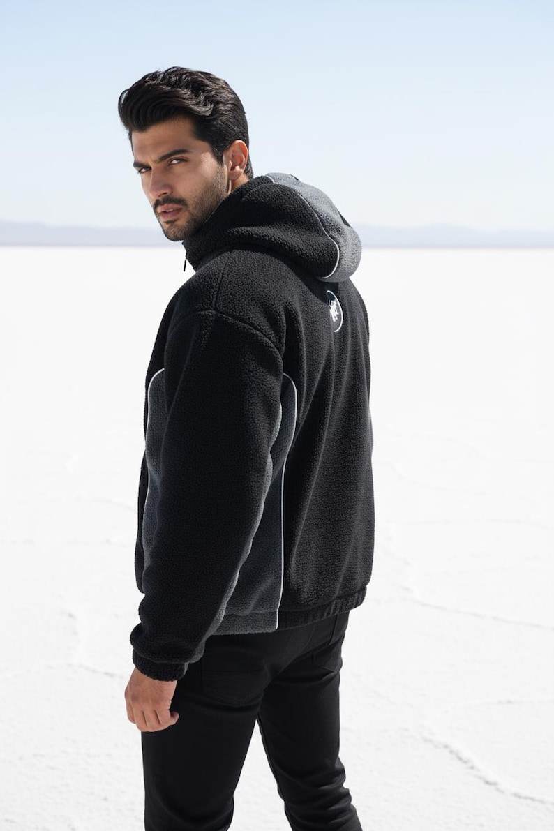 May include: Black fleece hooded jacket with a grey hood and white trim. The jacket has a zip closure and a small logo on the back. The model is wearing black trousers.