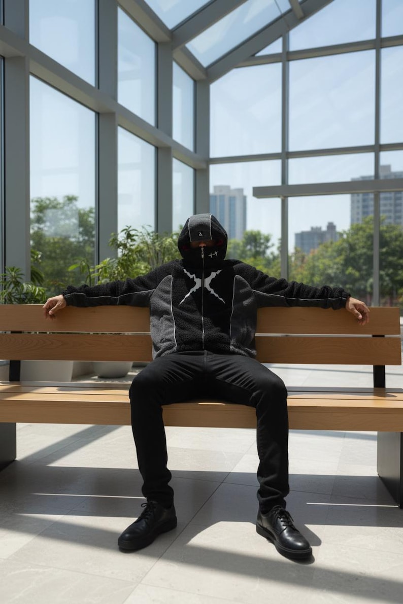 May include: A person wearing a black and grey hooded jacket with a white graphic design, black trousers, and black shoes, sitting on a wooden bench. The jacket has a zip and a hood that covers the head. The background includes large windows and a view of buildings.