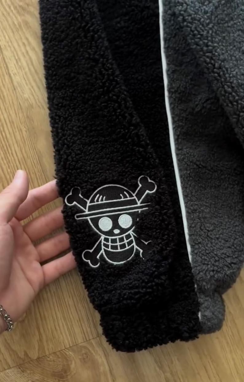 May include: Black and grey fleece jacket with white trim. The jacket has a white embroidered Jolly Roger patch on the sleeve. The jacket is made of a soft, textured material.
