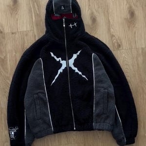 May include: Black fleece zip-up hoodie with a white graphic on the front. The hoodie features grey panels on the sleeves and a white stripe. The hood has a built-in face covering and a small logo. A pirate flag emblem is on the left sleeve.