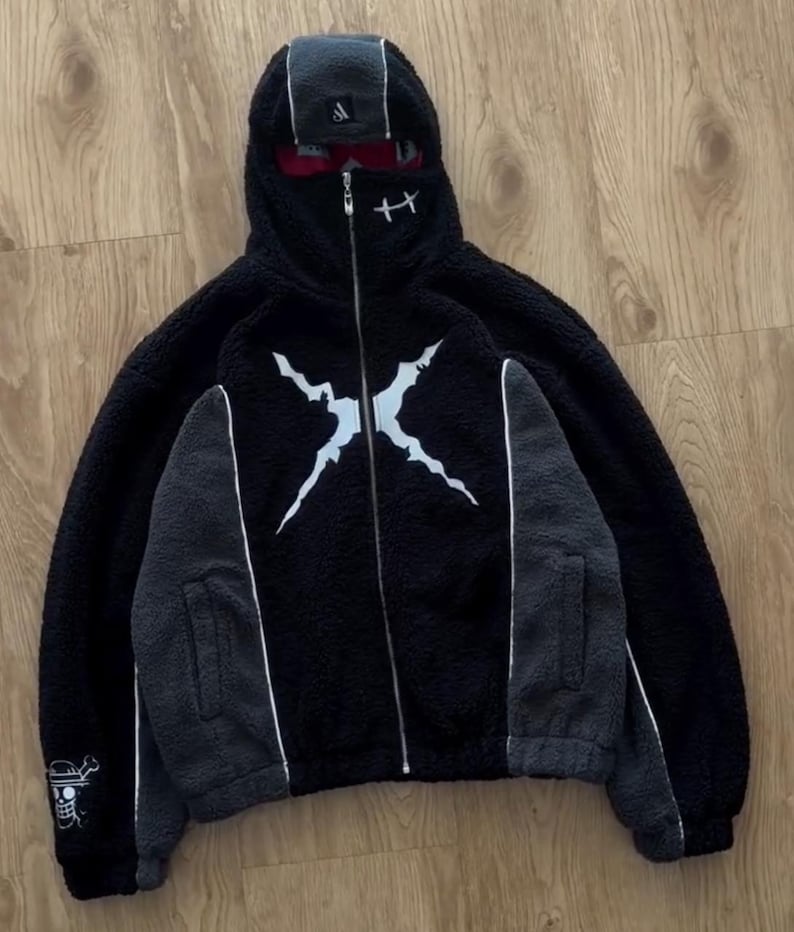 May include: Black fleece zip-up hoodie with a white graphic on the front. The hoodie features grey panels on the sleeves and a white stripe. The hood has a built-in face covering and a small logo. A pirate flag emblem is on the left sleeve.