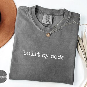 May include: A charcoal gray short-sleeve t-shirt with the text "built by code" printed in white on the chest. The shirt features a crew neckline and a Comfort Colors brand label on the inside collar, indicating a size small.