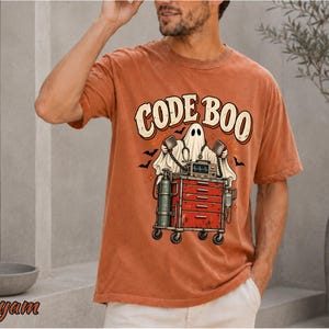 May include: A rust-colored short-sleeve t-shirt featuring a graphic of a ghost holding defibrillator paddles next to a red medical crash cart. The text CODE BOO is printed in a curved font above the ghost, accompanied by small black bats.