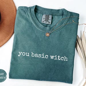 May include: A blue-green short-sleeve t-shirt with the white text you basic witch printed on the front. The shirt features a crew neckline and a Comfort Colors brand label on the inside collar. A gold-toned necklace rests on the fabric.