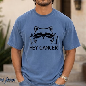 May include: A blue jean colored short-sleeve t-shirt featuring a black graphic of a cat wearing a mask and giving the middle finger with both paws. Below the cat, the text HEY CANCER is printed in a bold, black, sans-serif font.