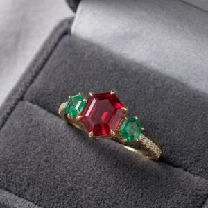 Pode incluir: Um anel de ouro com uma grande pedra preciosa vermelha central de corte hexagonal, ladeada por duas pedras verdes menores de corte hexagonal. A banda é fina e metálica, com pequenas pedras redondas transparentes ao longo do aro.