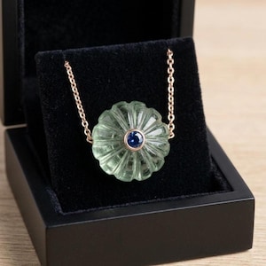 May include: A necklace with a light green, flower-shaped pendant and a dark blue central stone, suspended from a gold-coloured chain. The necklace is displayed in a black jewellery box.