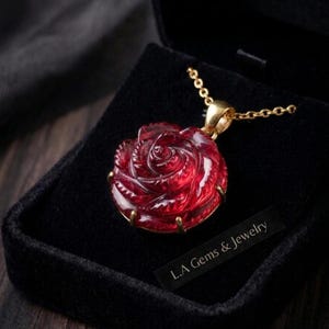 Puede incluir: Un colgante de piedra preciosa de color rojo intenso tallado con forma de rosa en flor, montado en un engaste de metal dorado con garras. El colgante cuelga de una cadena dorada a juego. Se ve una etiqueta negra con el texto L.A Gems & Jewelry.