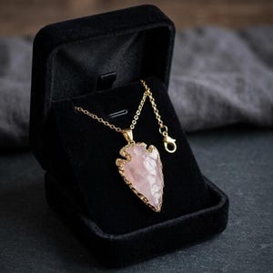 May include: A gold-colored necklace with a pendant shaped like an arrowhead, featuring a light pink gemstone. The necklace is displayed in a black velvet jewelry box, with a dark background.