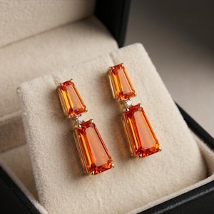 May include: A pair of drop earrings featuring two faceted orange gemstones in a tapered baguette shape, connected by a small clear stone set in metal. The earrings are displayed on a light beige fabric insert within a dark jewellery box.