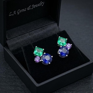 May include: A pair of stud earrings in a black L.A. Gems & Jewelry box. Each earring features a square-cut green gemstone, a round-cut blue gemstone, and a small round-cut purple gemstone set in silver-toned metal against a black velvet lining.