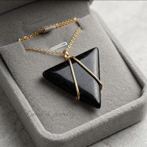 Può includere: Collana con pendente triangolare nero, con filo e catena dorati. La collana è esposta in un portagioie grigio. Il testo "L.A. Gems & Jewelry" è visibile.