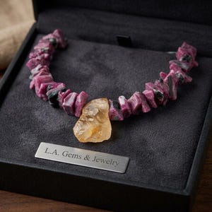 May include: A necklace of irregular, pink and green gemstones with a large, yellow-orange stone pendant. The necklace is displayed in a black jewelry box with a silver plate that reads "L.A. Gems & Jewelry."