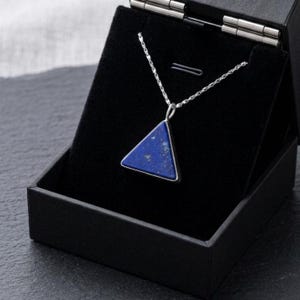 Op de afbeelding: Een zilveren ketting met een driehoekige, diepblauwe hanger, gepresenteerd in een zwarte sieradendoos. De hanger heeft een zilveren rand en een getextureerd oppervlak. De doos is open en onthult de ketting tegen een zwart fluwelen interieur.