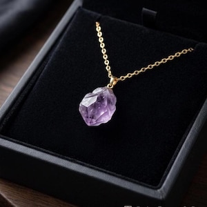 May include: A gold chain necklace with a light purple amethyst pendant. The gemstone is faceted and set in a gold bezel. The necklace is presented in a black jewelry box. A beautiful piece of jewellery.