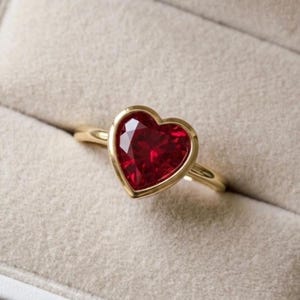 May include: A gold ring featuring a prominent heart-shaped red gemstone set in a smooth bezel setting. The band is thin and polished, showcasing a vibrant crimson stone with multiple facets that reflect light across the surface of the heart.