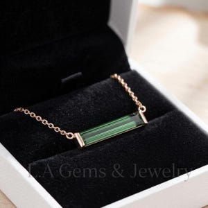 May include: A rose gold chain necklace featuring a horizontal, rectangular, green tourmaline baguette gemstone set in a metal frame. The jewelry is displayed inside a black velvet-lined box with the text L.A Gems & Jewelry printed on the base.