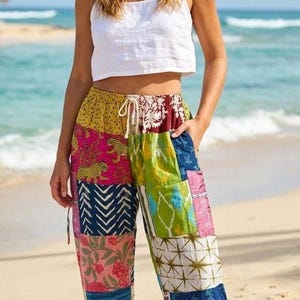 Peut inclure: Une personne porte un haut court blanc et un pantalon patchwork coloré sur une plage. Le pantalon présente divers motifs et couleurs, notamment rose, jaune, bleu et vert. Le pantalon a une taille à cordon.