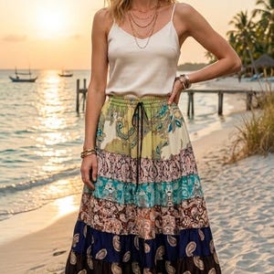Puede incluir: Una mujer con una camiseta sin mangas color crema y una falda larga escalonada con estampado de cachemira y floral. La falda tiene cintura con cordón y varios colores, como azul, marrón y verde. El fondo es una escena de playa.