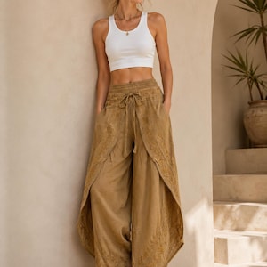 Könnte beinhalten: Eine Frau trägt ein weißes geripptes Tanktop und eine beige bestickte Palazzo-Hose mit Kordelzug in der Taille. Die Hose hat ein geteiltes Frontdesign. Sie trägt Sonnenbrille und Sandalen. Der Hintergrund ist eine neutral getönte Wand und ein Torbogen.