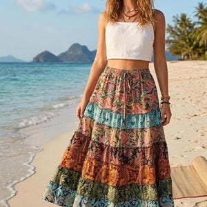 Könnte beinhalten: Eine Frau trägt ein weißes Crop-Top und einen langen, gestuften Rock mit einem farbenfrohen Paisley-Muster. Der Rock zeigt Farbtöne von Rosa, Blau, Orange und Grün. Die Szene spielt sich an einem Sandstrand mit dem Meer im Hintergrund ab.