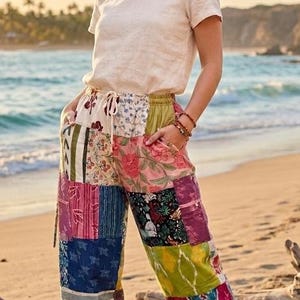 May include: A woman wearing a cream-colored t-shirt and patchwork pants on a beach. The pants feature a variety of colorful fabric patterns, including floral and geometric designs. The pants have a drawstring waist and are cuffed at the ankles. The woman is wearing brown sandals.