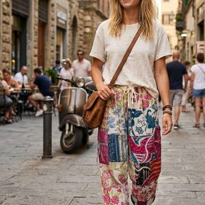 May include: A woman wearing a cream-colored t-shirt and patchwork pants with floral and geometric patterns. She carries a brown leather crossbody bag and wears white sneakers. The background shows a European street scene.