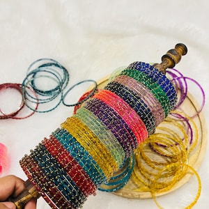 Op de afbeelding: Een verzameling dunne, doorschijnende armbanden in diverse kleuren zoals blauw, groen, roze, zwart, rood, geel en paars, allemaal voorzien van kleine goudkleurige spikkels. De armbanden zijn op een houten staaf gestapeld en verspreid.