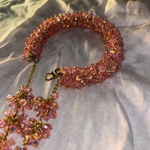 Könnte beinhalten: Ein rosa Perlenarmband mit einer goldenen Kette und einem Verschluss. Das Armband besteht aus kleinen, schillernden rosa Perlen. Eine goldene Kette mit rosa Perlenclustern hängt am Armband.
