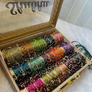 May include: A collection of multi-colored metal bangles arranged in a rectangular storage box with a clear lid. The bangles feature a thin gold-tone wire wrap with small decorative beads and come in shades of pink, green, gold, blue, and purple.