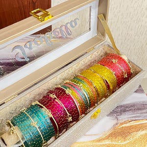 May include: A rectangular beige storage box with a clear lid containing a row of multi-colored bangles. The bangles are arranged in a gradient of teal, magenta, pink, green, yellow, and red, each featuring small gold-toned accents and beads.