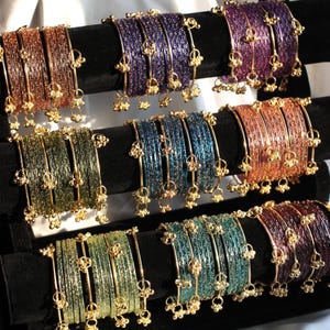 Puede incluir: Exhibición de pulseras coloridas en tonos marrón, morado, azul, verde y naranja. Cada pulsera está adornada con detalles dorados y pequeños dijes colgantes. Las pulseras están dispuestas sobre soportes de terciopelo negro, mostrando sus intrincados diseños.